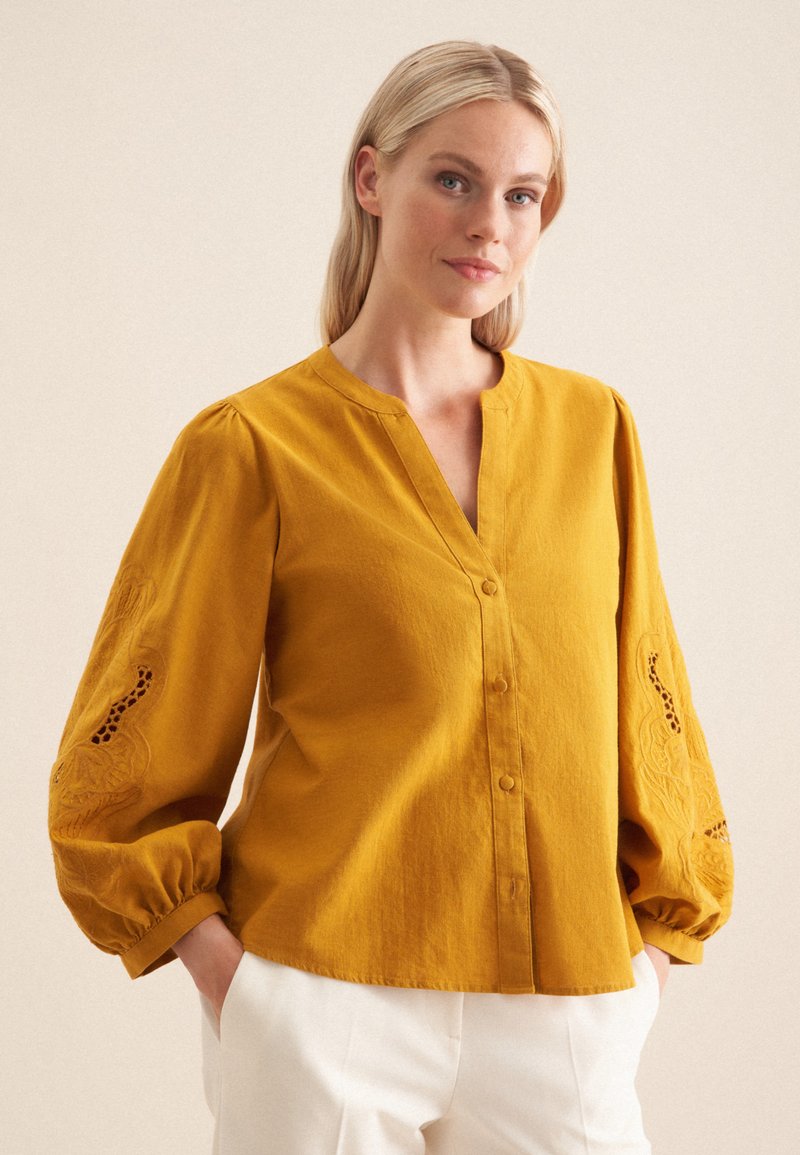 Camicia button-up gialla senape con scollatura a V, maniche a palloncino e dettagli ricamati sulle maniche. Tessuto morbido con una finitura testurizzata.
