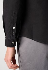 Brazo izquierdo con una camisa de manga larga negra con botones blancos, junto a pantalones gris claro, mostrando detalles del puño de la camisa y la costura lateral.