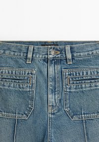Modré džíny z denimu s předními klopovými kapsami, poutky na opasek a kovovým knoflíkem na pase, zobrazené z přední oblasti pasu.