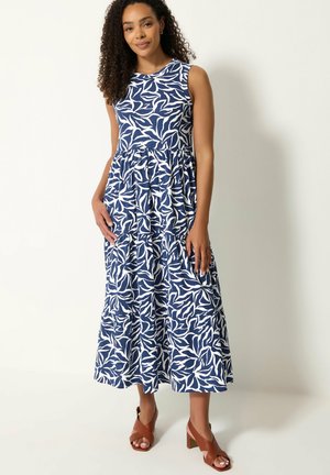 Mujer con vestido midi sin mangas con estampado azul y blanco, y sandalias marrones de tacón, de pie frente a un fondo liso y claro.
