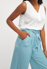 Haut blanc sans manches avec un col en V et une poche, associé à un pantalon large bleu clair à taille nouée et poches latérales.