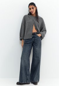 Žena v šedém kardiganu se zapnutým jedním knoflíkem, širokých tmavých džínách, černém pásku a černých botách, stojící na bílém pozadí.