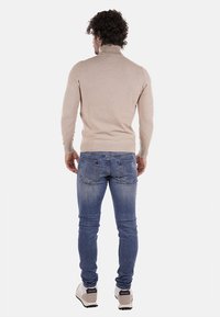 Maglione beige a maniche lunghe con colletto a lupetto realizzato in morbido tessuto a maglia, abbinato a jeans blu slim-fit, visto da dietro.