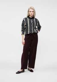 Šviesiaplaukė moteris stovi su ranka kišenėje, vilkinti juodai ir smėlio spalvos deimantų rašto megztinį, tamsias plačias kelnes ir bordo spalvos butus.