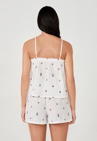 Kobieta ubrana w białą koszulkę na ramiączkach i szorty w małe różowe kwiatowe wzory, pokazana z tyłu na gładkim jasnym tle.