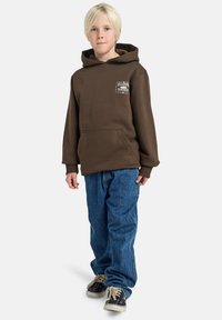 Brun hettegenser med frontlomme, kombinert med blå denimjeans. Har svart og hvit grafisk detalj på brystet.