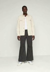 Camisa bege clara de botões, t-shirt branca, jeans largos escuros, sapatilhas brancas com detalhes pretos. Modelo está encostado a um fundo simples.