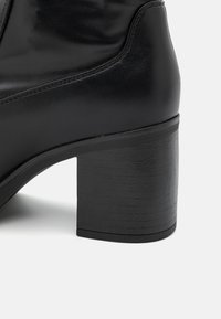 Botte de cheville noire à talons, en cuir lisse. Présente un talon épais à texture et des coutures discrètes le long de l'empeigne.