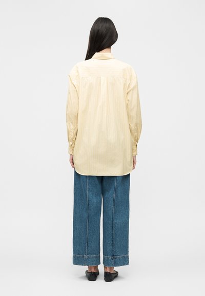 Γυναίκα με μακριά σκουρόχρωμα μαλλιά φοράει ένα φαρδύ ανοιχτό κίτρινο πουκάμισο με μακριά μανίκια, φαρδιά μπλε τζιν και μαύρα επίπεδα παπούτσια, φαίνεται από πίσω.