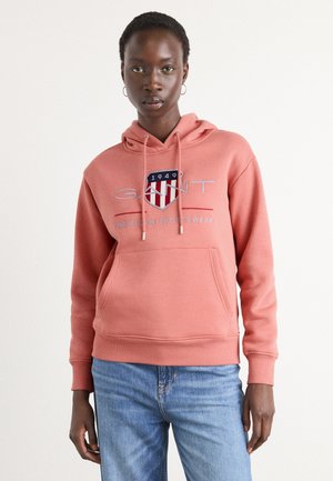 Koral fleece hettegenser med frontlomme, snor i hetten og brodert logo. Kombinert med blå denimjeans. Enkel, uformell design.