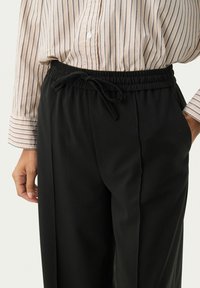 Pantalons noirs avec une taille élastique et un cordon de serrage, dotés de poches latérales et d'une texture lisse, associés à une chemise rayée à boutons.