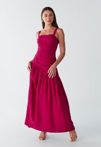 Vestido largo fucsia con corpiño ajustado fruncido con tirantes finos y falda fluida y recogida. Tela suave con un ligero brillo.