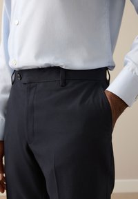 Homme portant un pantalon noir habillé et une chemise bleu clair boutonnée, une main dans la poche.