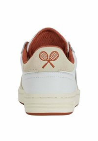 Zapatilla blanca con una lengüeta de ante marrón, forro de malla roja y logo de raqueta de tenis naranja en la parte posterior. Suela de goma con rayas horizontales.