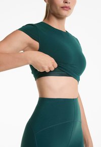 Žena podiže tamnozeleni kratki crop top, otkrivajući elastični pojas sportskog grudnjaka, nosi odgovarajuće tajice viskog struka.