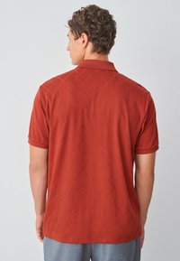 Polo shirt de couleur argile foncée avec un motif de diamant texturé, à manches courtes et col; le tissu est lisse et légèrement extensible.