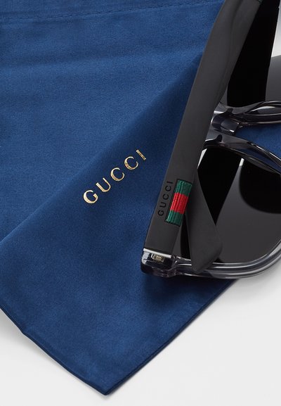Sluneční brýle Gucci odložené na texturované tmavě modré látce; mají černé čočky, šedé ramena a detail loga se zeleno-červenou-zelenou pruhovanou linkou.
