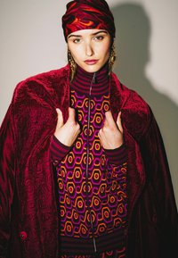 Cappotto in velluto bordeaux con motivo testurizzato, maglione aderente con disegni esagonali in viola, arancione e giallo; foulard rosso con motivo.