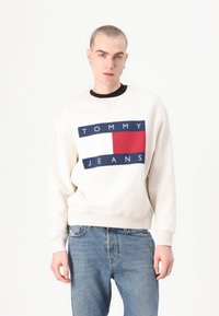 Krémová mikina s grafikou „TOMMY JEANS“ v námorníckej modrej a červenej s obdlžníkovými akcentami. V kombinácii so svetlomodrými denimovými džínami.