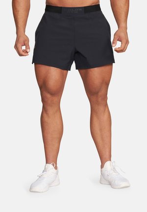 Umięśniona osoba w czarnych sportowych szortach i białych tenisówkach, stojąca z rękami swobodnie opuszczonymi wzdłuż ciała na jednolitym tle.