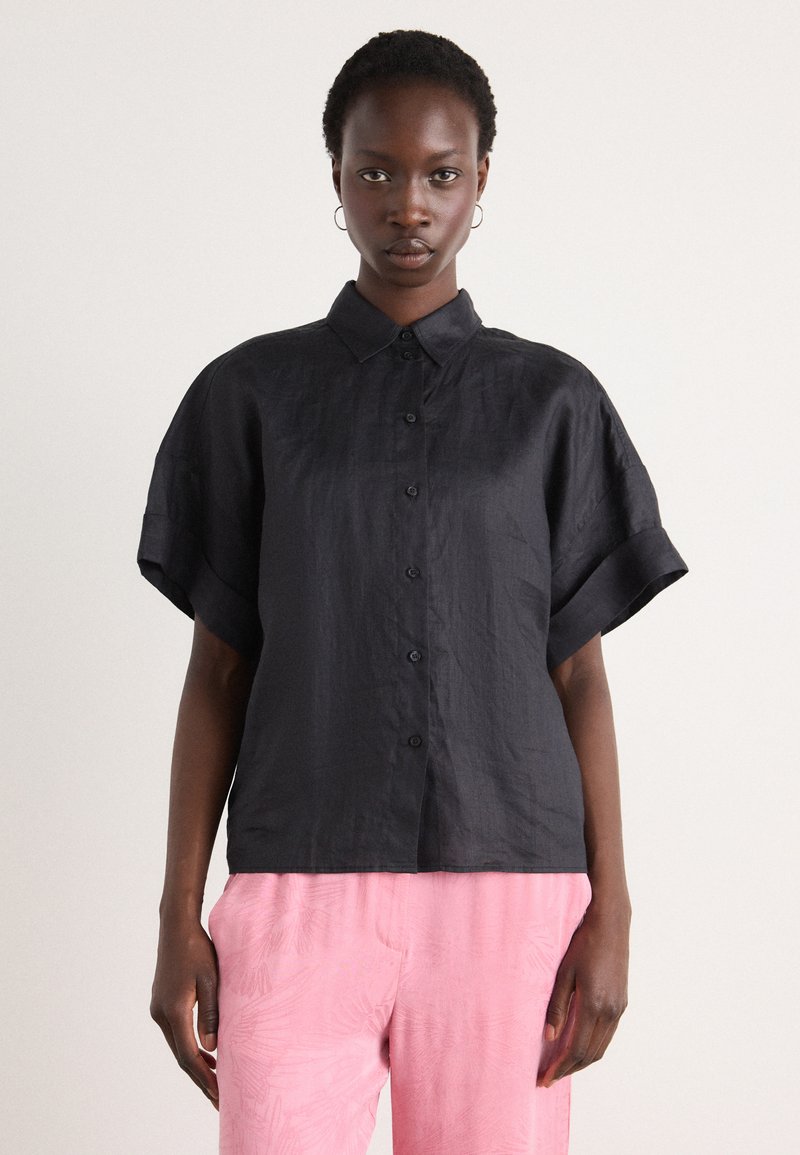 Camicia nera a maniche corte con bottoni realizzata in tessuto testurizzato. Presenta un colletto e maniche arrotolate, abbinata a pantaloni rosa con motivo.