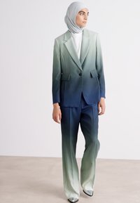 Zweiteiliger Anzug in grünen Farbverläufen, bestehend aus einem Blazer mit Revers und passenden Hosen, aus glattem Stoff. Silberne Schuhe.