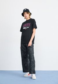 Black t-shirt with pink Nike logo, patterned black wide-leg pants, and white sneakers with red accents. Black bucket hat. Solid background.