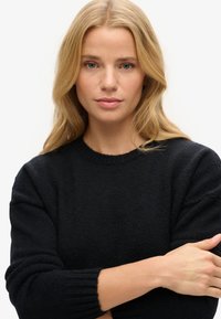 Czarny sweter dzianinowy z okrągłym dekoltem, prążkowanymi mankietami i miękką fakturą. Model ma skrzyżowane ramiona, prezentując dopasowanie ubrania.