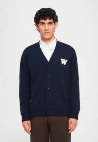 Námornícky pletený cardigan s V-výstrihom, šiestimi čiernymi gombíkmi, rebrovanými manžetami, so zeleným vyšitým logom "W" na ľavej hrudi.