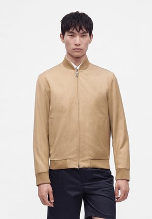Homme portant une veste beige zippée et un short foncé, debout devant un fond blanc uni, face à la caméra avec une expression neutre.