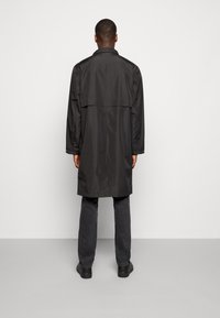 Gabán impermeable negro de corte recto, con mangas largas y un detalle de ventilación en la parte posterior. Lleva puesto sobre unos jeans oscuros y zapatos negros.