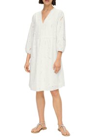 Robe blanche à mi-longueur avec un décolleté en V, une broderie décorative et de courtes manches bouffantes. Portée avec de simples sandales à lanières.