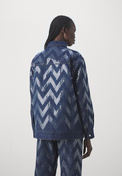 Denim-jakk, millel on hõbedast sequinidega chevron-muster sinise taustaga. Tugev ja struktureeritud disain, millel on nööpdetailid.