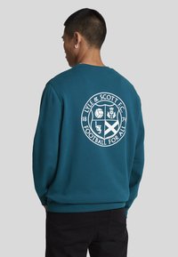 Sudadera verde azulado con un gráfico redondo blanco en la parte posterior que presenta "Lyle & Scott F.C." y varios símbolos de fútbol. Textura suave, corte relajado.