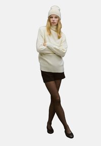 Kremowy sweter z golfem z prążkowanym obszyciem, zestawiony z czarnymi szortami. Modelka nosi jasnobeżową czapkę typu beanie i czarne baleriny, a także cienkie czarne rajstopy.