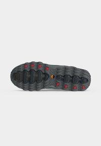 Semelle de chaussure noire avec des rainures profondes et des grips circulaires rouges, avec un petit logo circulaire orange au centre sur un fond blanc.
