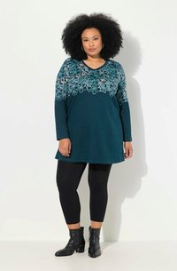 Teal pitkähihaisen tunika, jossa on valkoista kukkakirjontaa kaula-aukon ympärillä, yhdistetty mustiin legginsseihin ja nilkkureihin. Pehmeä kangas.