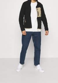 Czarna polarowa kurtka z beżowymi akcentami i kieszeniami na zamek, noszona na białym t-shircie i niebieskich dżinsach, w połączeniu z białymi trampkami.