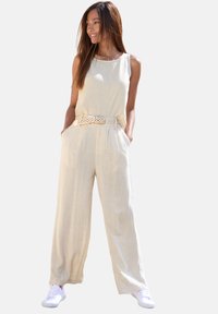 Light beige linen jumpsuit with wide legs, sleeveless design, equipped with a textured white woven belt, paired with white sneakers.