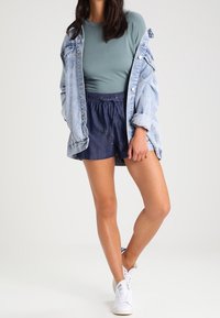 Un short en denim bleu foncé, assorti à un haut à manches longues bleu clair et une veste en denim oversized clair. Des baskets blanches complètent la tenue.