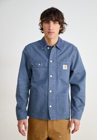Blå denim arbeidsjakke med knappestolpe, to brystlommer og to nedre lommer. Har messingknapper og en klassisk kragedesign.