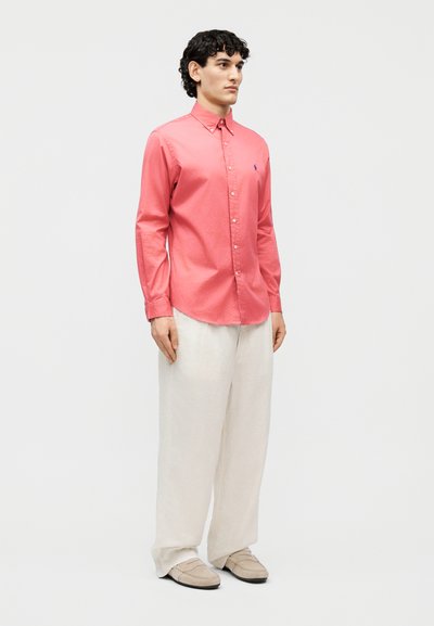 Άνδρας που στέκεται με λευκό, άνετο παντελόνι, μπεζ μοκασίνια και ροζ πουκάμισο με κουμπιά και μακριά μανίκια, μπροστά σε απλό φόντο.