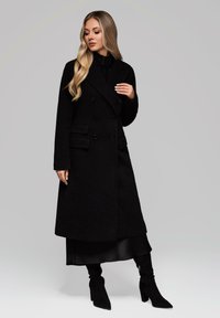 Cappotto nero doppiopetto con tasche laterali, realizzato in un tessuto testurizzato, caratterizzato da una silhouette lunga e dritta e grandi bottoni.