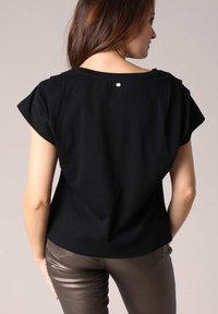 Zwarte korte mouwen top met een losse drape, voorzien van een ronde halslijn en een kleine metaalaccent aan de achterkant. Gecombineerd met metallic afwerking broek.