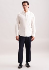 Een man staat in een witte, knoopsluiting, lange mouwen shirt, donkere marineblauwe broek en zwarte nette schoenen tegen een effen lichte achtergrond.