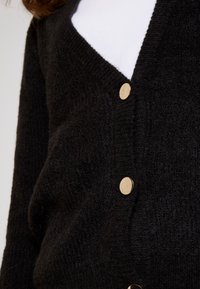 Black textured knit cardigan with gold round buttons worn over a white top, showing upper torso and part of brown hair.