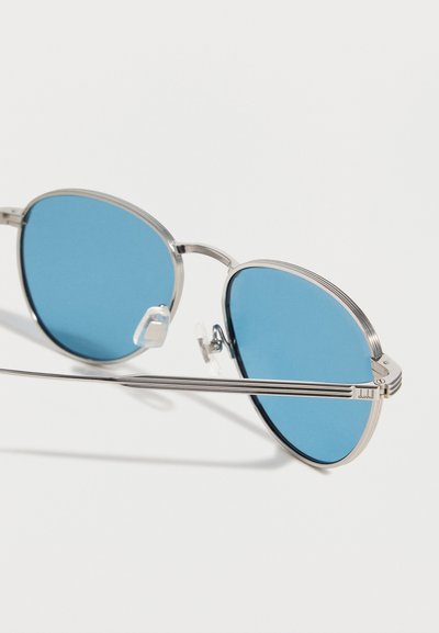 Γυαλιά ηλίου με ασημένιο μεταλλικό σκελετό, στρογγυλούς μπλε φακούς και ρυθμιζόμενα μαξιλαράκια μύτης που ακουμπούν σε λευκή επιφάνεια.