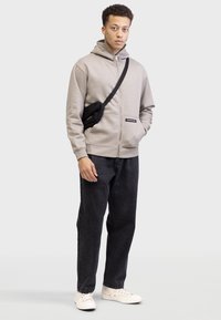 Sweat à zip gris clair avec un col haut, accents noirs, associé à un pantalon en denim foncé et des baskets blanches. Sac bandoulière noir porté.