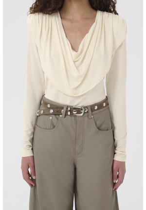 Femme portant un haut crème à manches longues avec un col drapé et un pantalon taille haute taupe avec une double ceinture cloutée argentée, debout, les bras relâchés.
