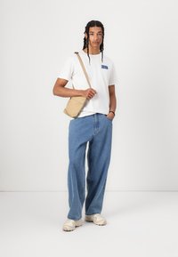 Lätt beige crossbody-väska, vit t-shirt med blå grafik, löst sittande blå denimjeans och neutrala sneakers. Enkel och avslappnad outfit.